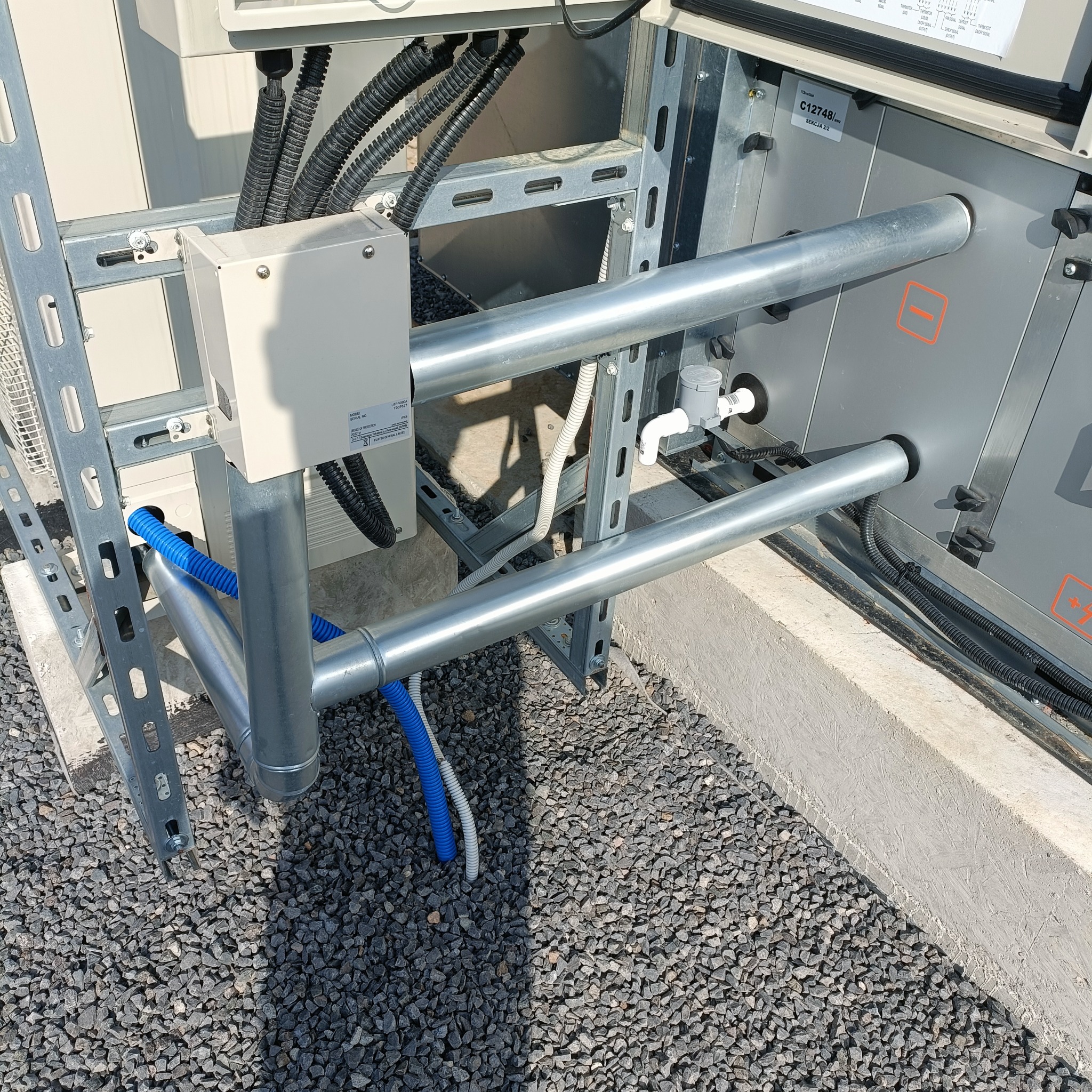 Szara instalacja z rurami i przewodami na żwirowym podłożu, widoczne elementy montażowe i skrzynka elektryczna. Detal instalacji klimatyzacji lub wentylacji.