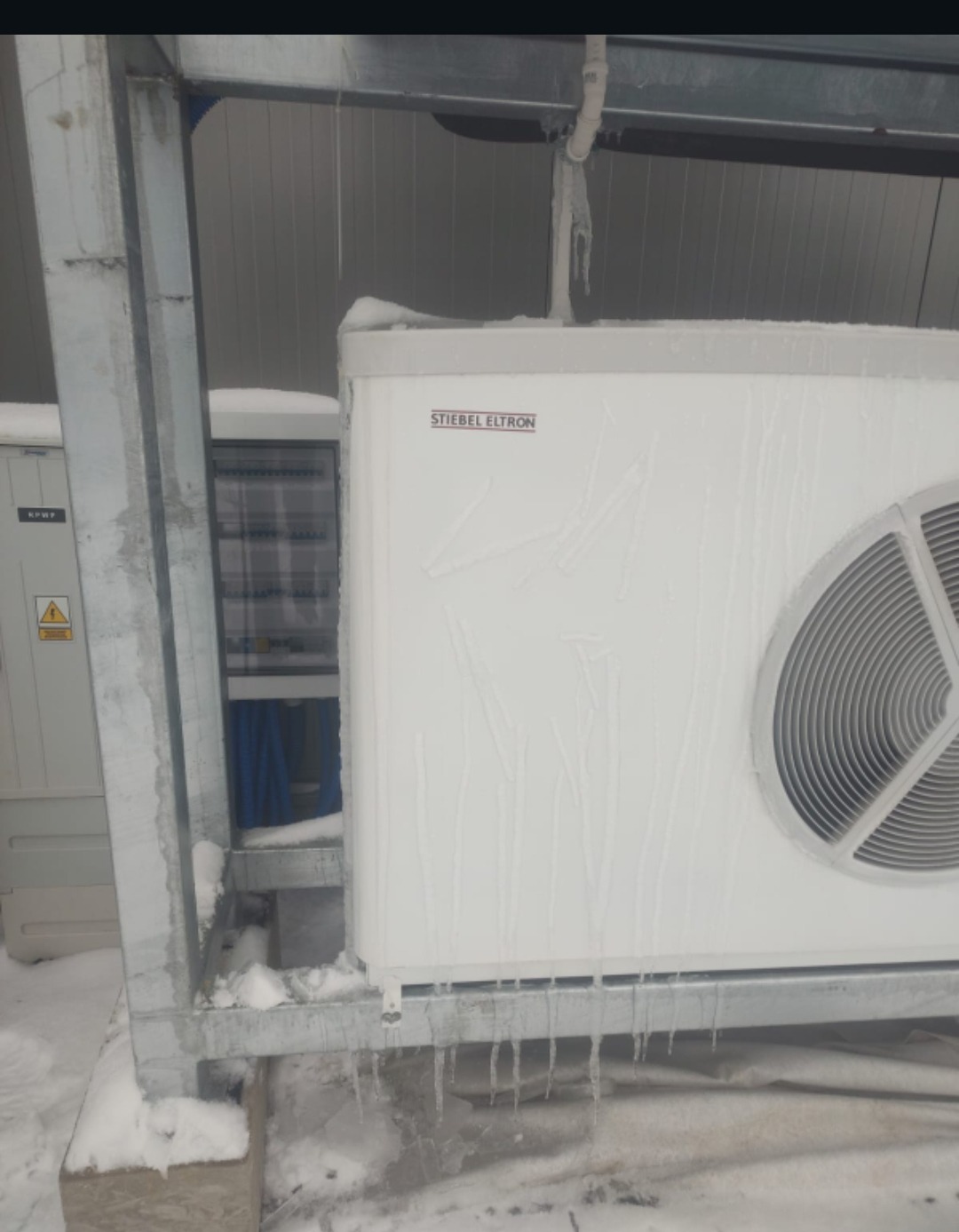 Biała pompa ciepła Stiebel Eltron, pokryta śniegiem i lodem, z widocznymi soplami lodu. Metalowa konstrukcja wsporcza. Zimowa sceneria.
