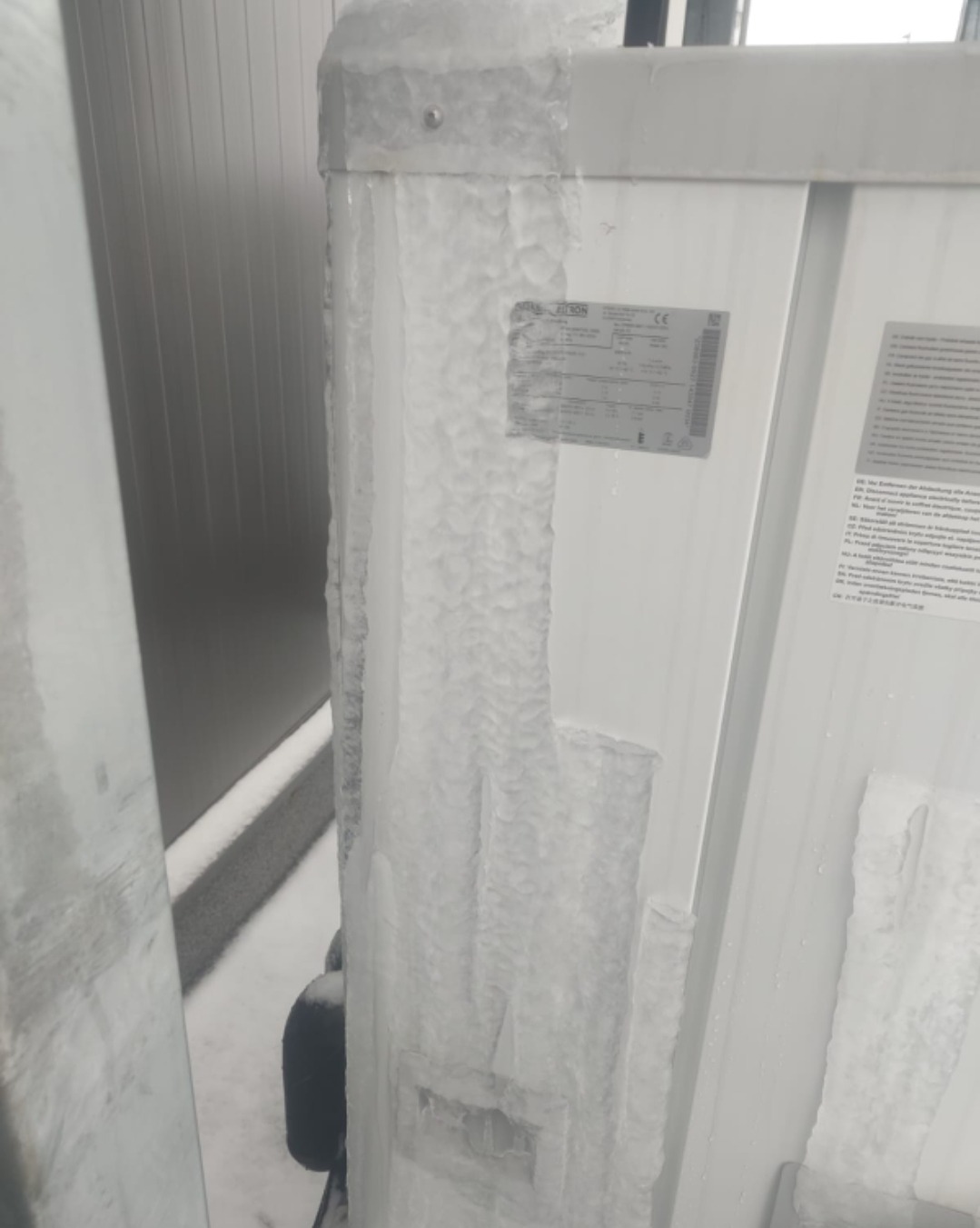Zamrożona obudowa pompy ciepła na zewnątrz budynku, pokryta grubą warstwą lodu. Widoczne tabliczki znamionowe i fragment otoczenia w zimowej scenerii.