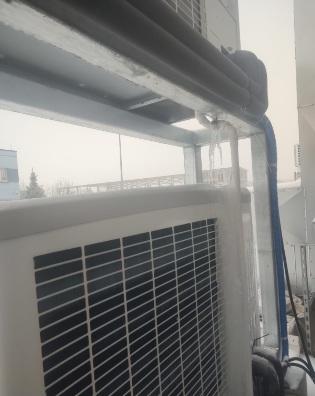 Zamrożona klimatyzacja zewnętrzna z widocznymi lodowymi sopelkami i oszronionymi rurami, na tle pochmurnego nieba i budynku w oddali. Widoczne elementy konstrukcji wsporczej.