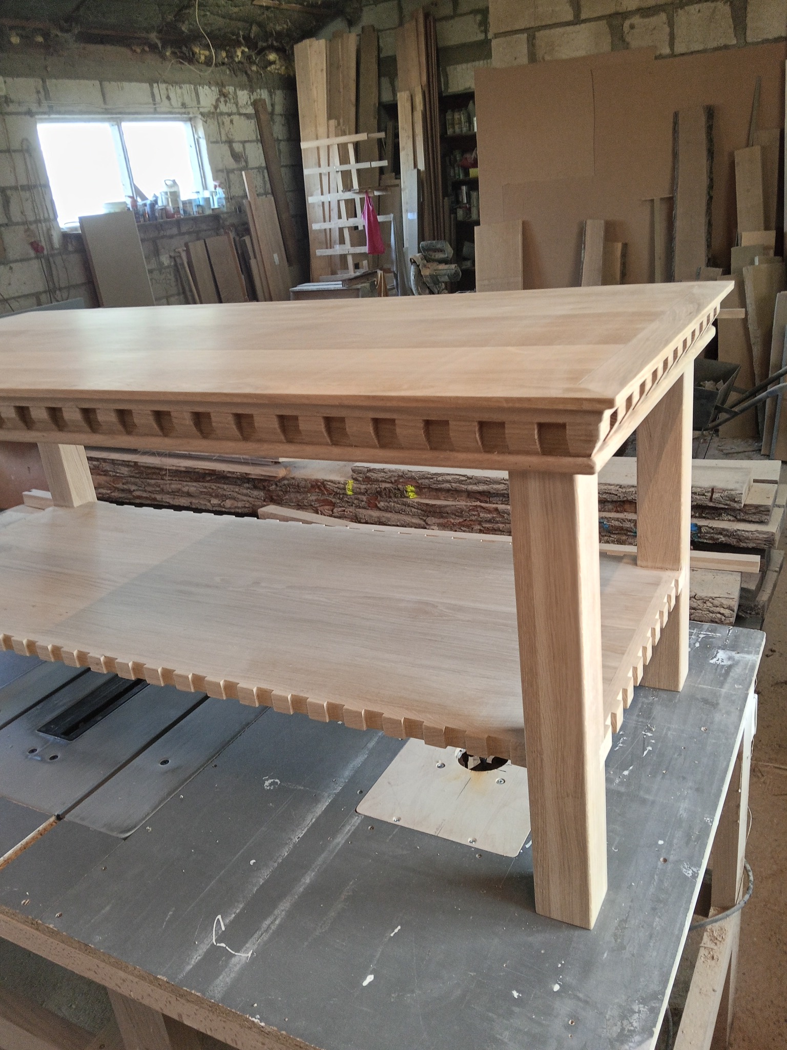 Surowy, drewniany stolik kawowy z dekoracyjnymi wycięciami, stojący na stole warsztatowym w pracowni stolarskiej. Widoczne deski i narzędzia w tle.