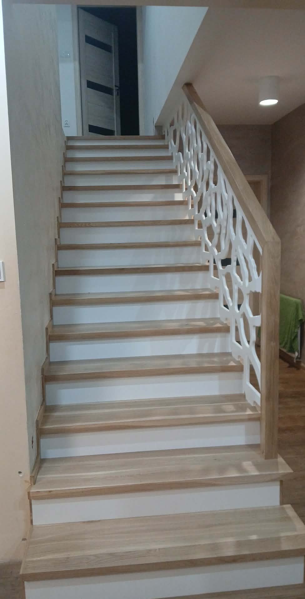 Jasne, drewniane schody z białymi podstopnicami i ażurową balustradą. Widok z dołu, podkreślający detale wykończenia i nowoczesny design.