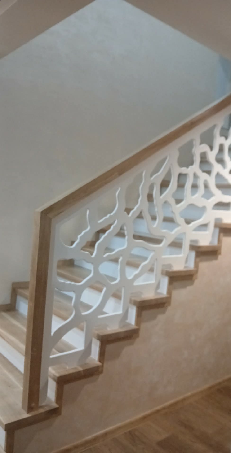 Drewniane schody z ażurową, białą balustradą o organicznym wzorze, widoczne z boku. Stopnie schodów w jasnym odcieniu drewna, ściana w tle beżowa.