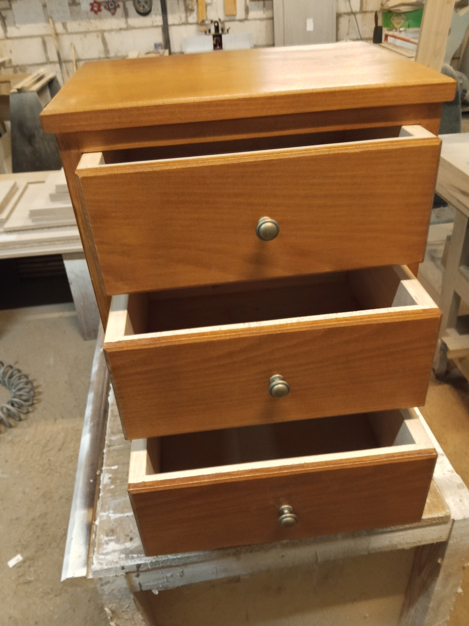 Drewniana komoda z trzema wysuniętymi szufladami, widoczne wnętrza szuflad. Wykończenie w ciepłym odcieniu brązu, okrągłe uchwyty. Stoi na roboczym stole w warsztacie.