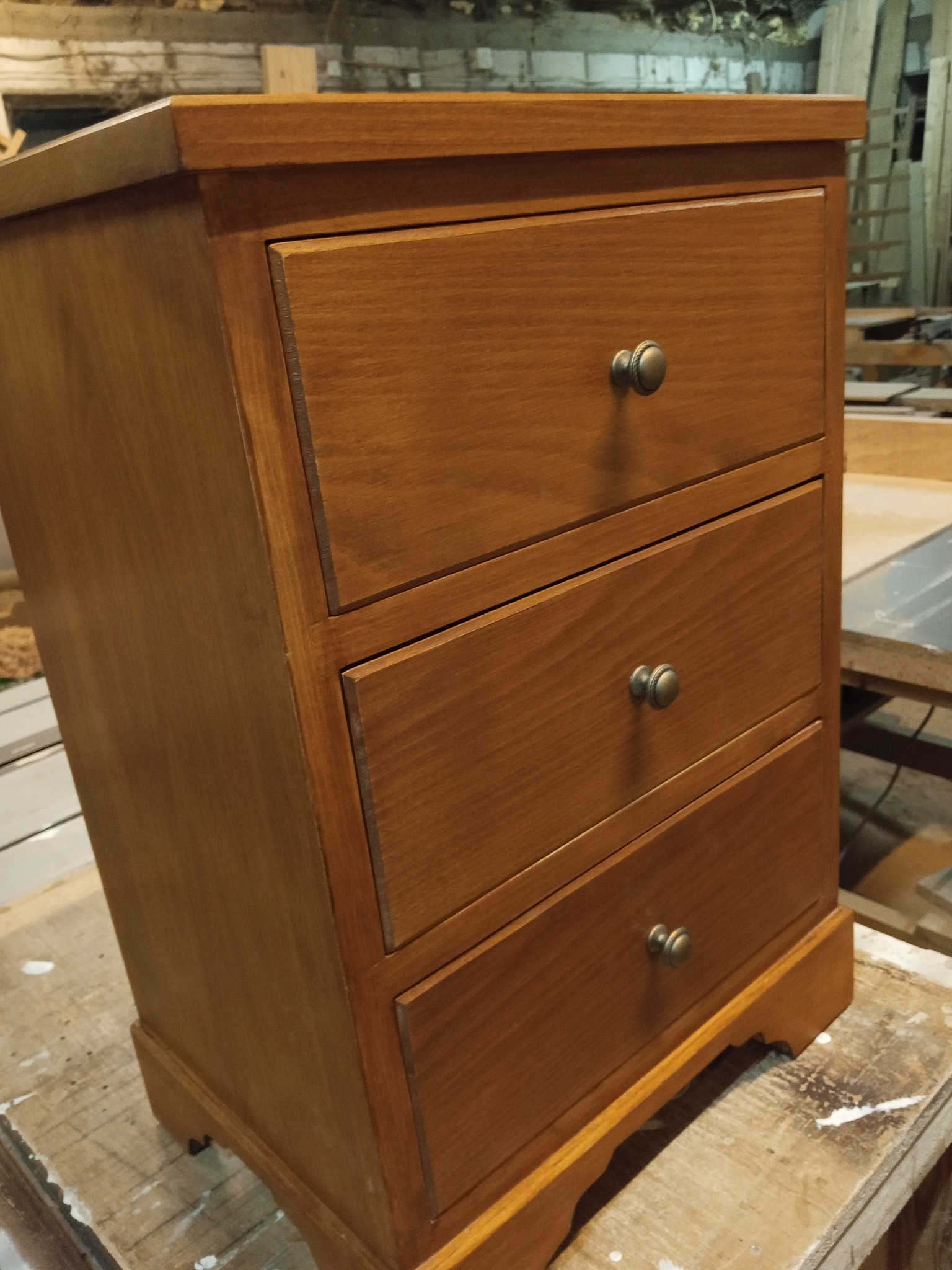 Drewniana komoda z trzema szufladami, wykończona w ciepłym odcieniu brązu, z metalowymi gałkami. Widoczne detale stolarskie i naturalne usłojenie drewna.