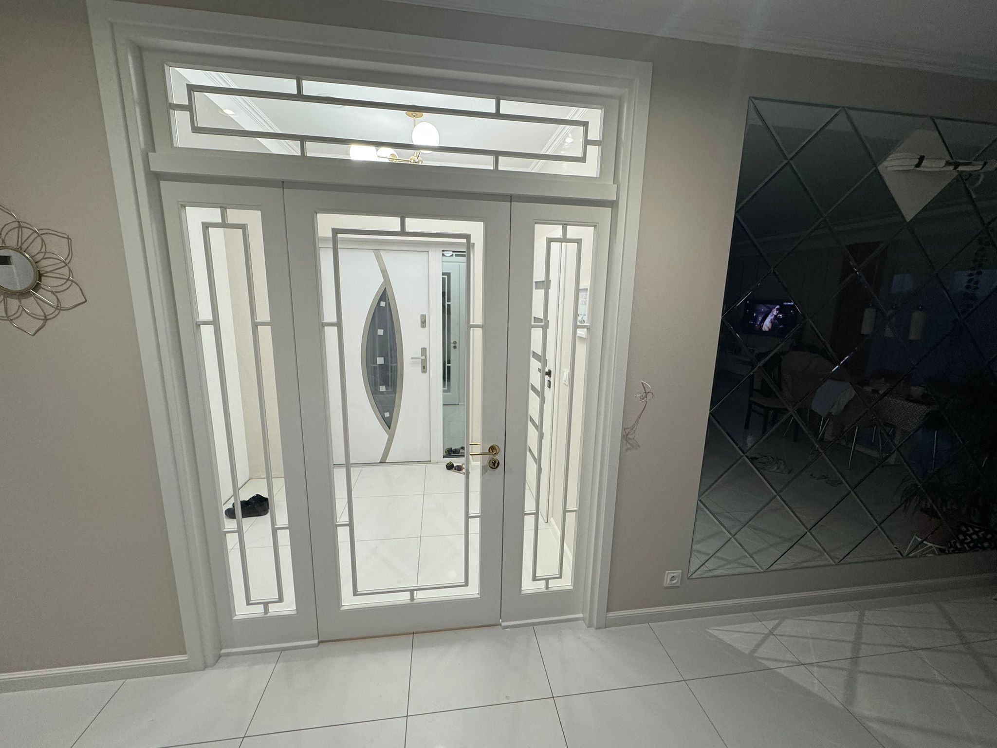 Białe, przeszklone drzwi wewnętrzne z nadświetlem, widok z bliska. W tle widoczny fragment lustrzanej ściany i podłoga z białych płytek.