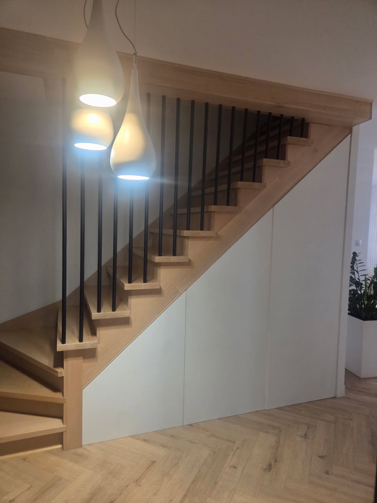 Drewniane schody z czarnymi balustradami i zabudową pod schodami w kolorze białym. Nad schodami wiszą trzy lampy w kształcie kropli.