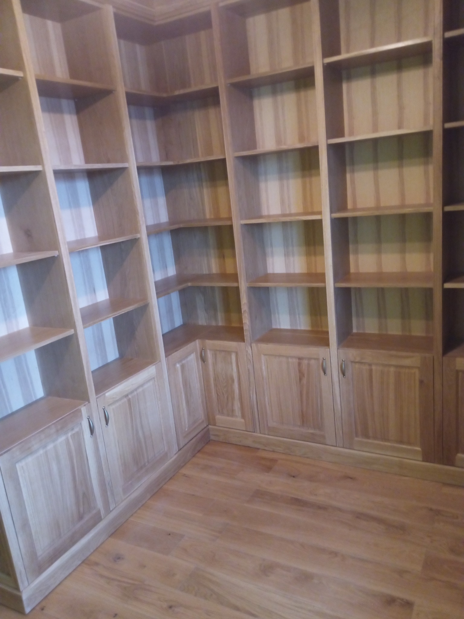 Drewniana zabudowa biblioteczna z szafkami i półkami, wykonana na wymiar, z widoczną strukturą drewna i dopasowaniem do kąta pomieszczenia.