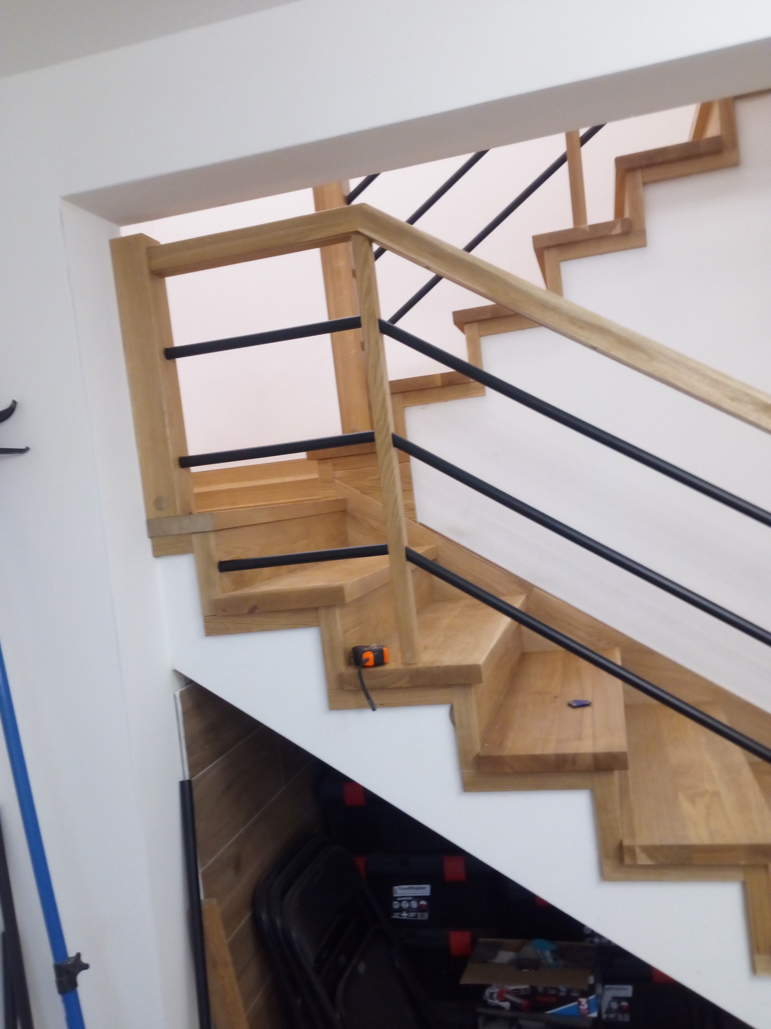 Drewniane schody z czarnymi, metalowymi balustradami, oświetlone naturalnym światłem, z widocznym miernikiem na jednym ze stopni. Minimalistyczny design.