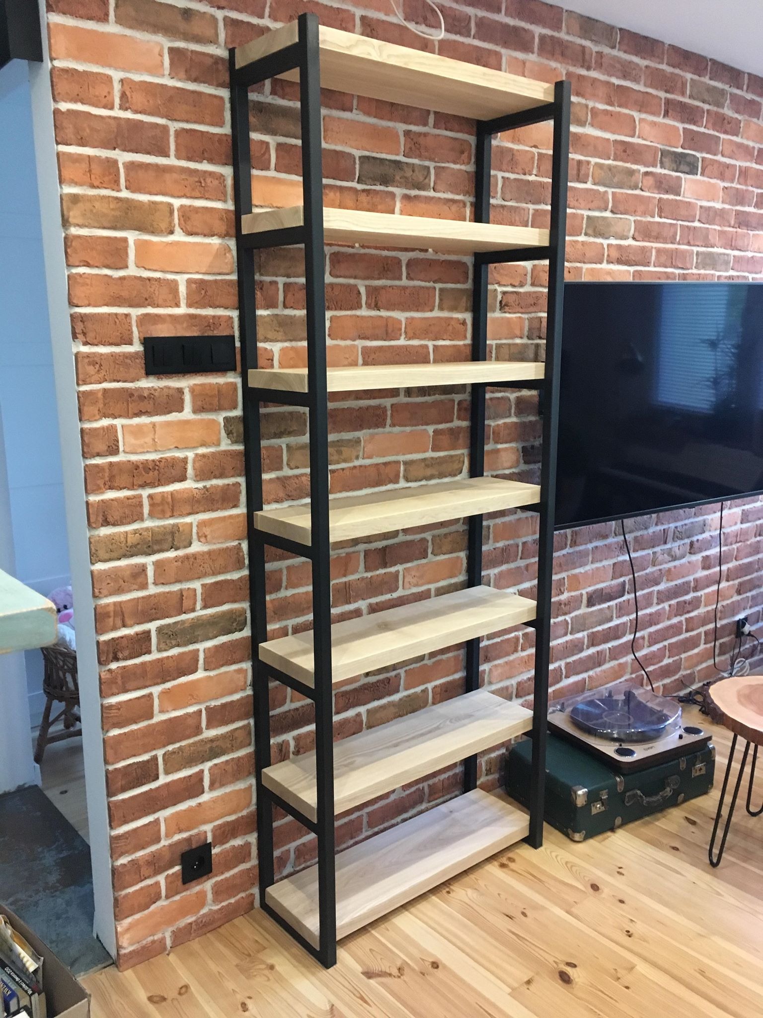 Regał loftowy z jasnego drewna i czarnego metalu na tle ściany z czerwonej cegły. Widoczny gramofon na walizce i drewniana podłoga.