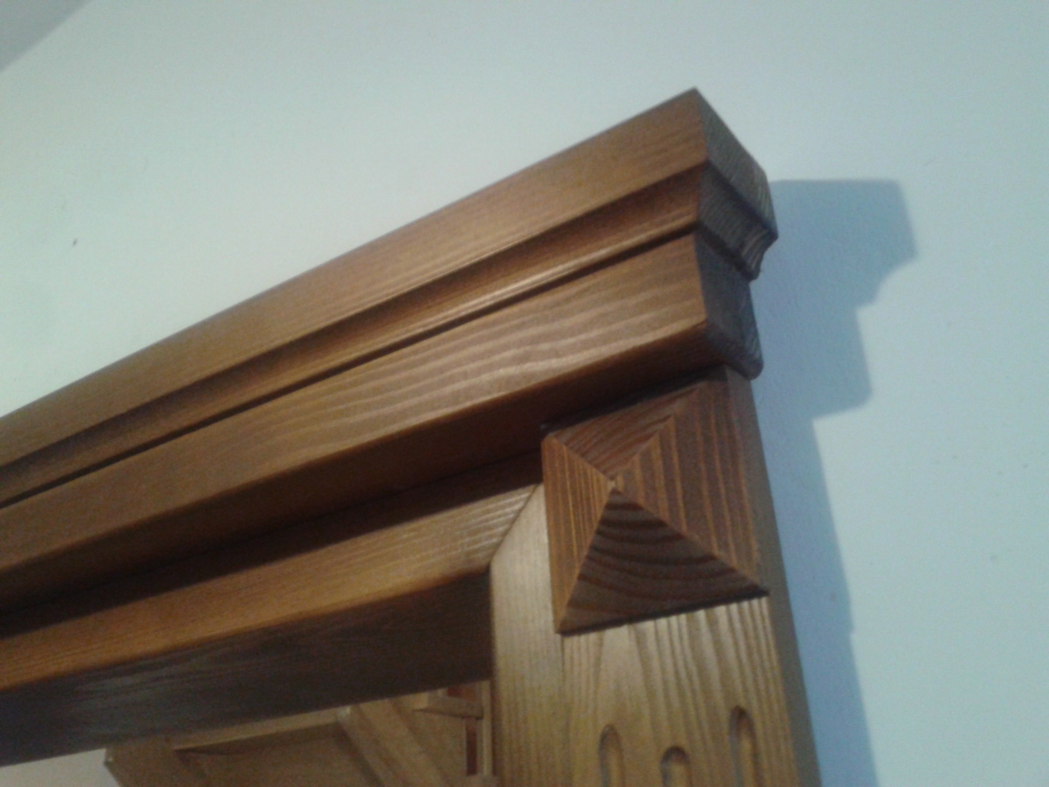 Fragment drewnianej ościeżnicy drzwiowej z dekoracyjnymi elementami, widoczny od góry pod kątem. Jasne drewno z wyraźnym usłojeniem, piramidalne zakończenie.