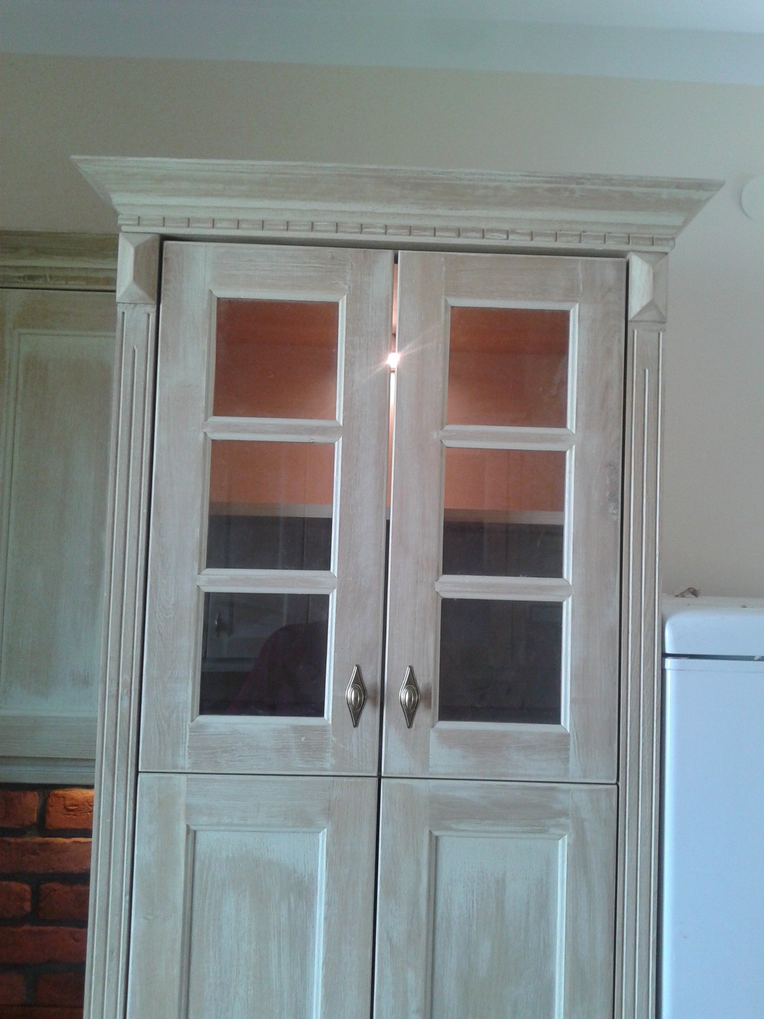 Dwudrzwiowa szafka kuchenna z przeszklonymi górnymi drzwiczkami, w stylu rustykalnym, z dekoracyjnym wykończeniem i metalowymi uchwytami. Widoczny fragment ceglanej ściany.