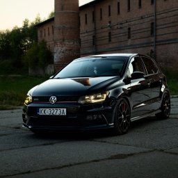 Dmitrowsky Content Lab - Czarne auto VW Polo GTI na tle ceglanego budynku z kominem. Reflektory włączone, tablica rejestracyjna widoczna. Zdjęcie wykonane o zmierzchu.