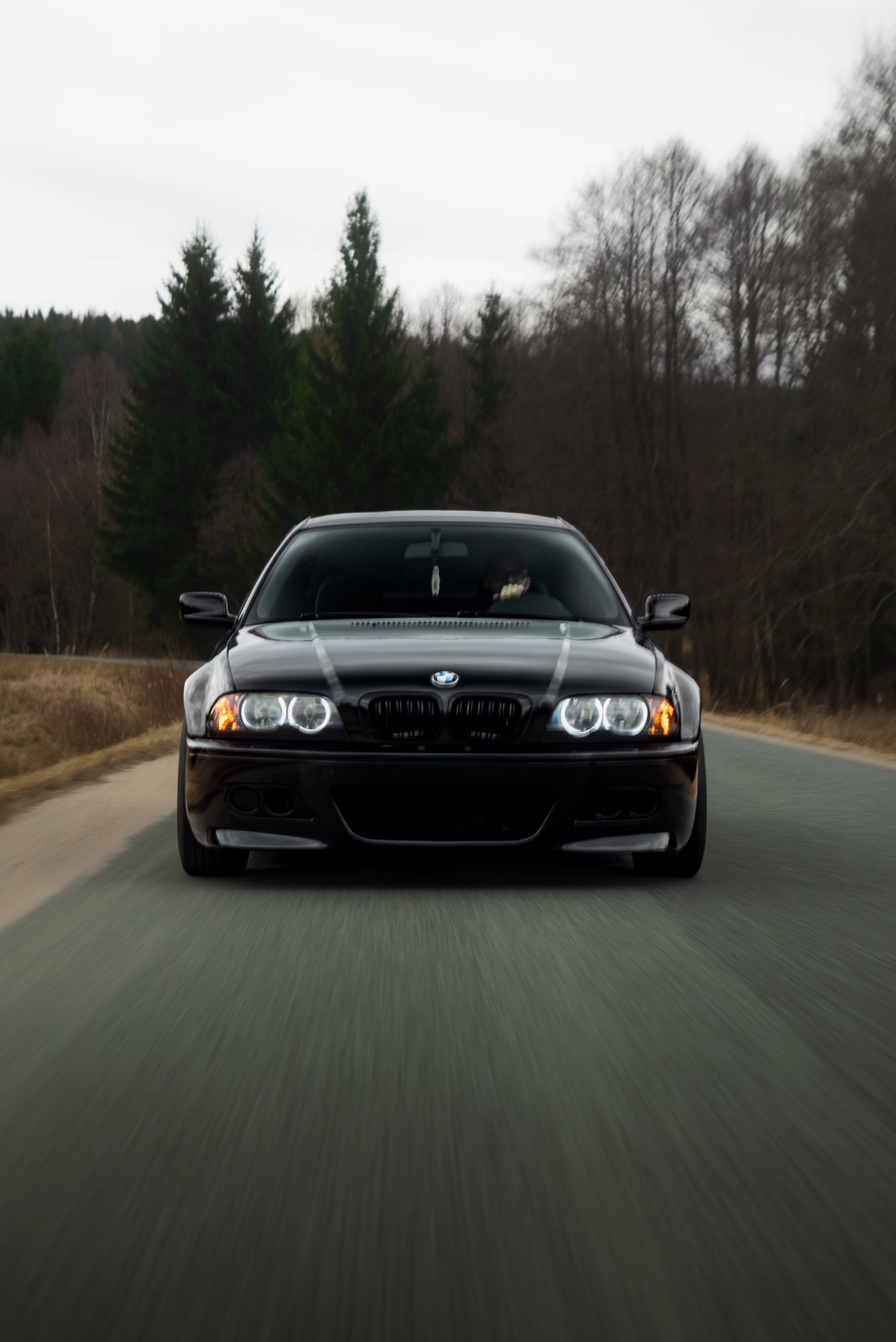 Dynamiczne ujęcie z przodu czarnego BMW E46 M3 na drodze, z rozmytym tłem drzew i nieba. Charakterystyczne ringi świateł i niski kąt kamery podkreślają sportowy charakter auta.