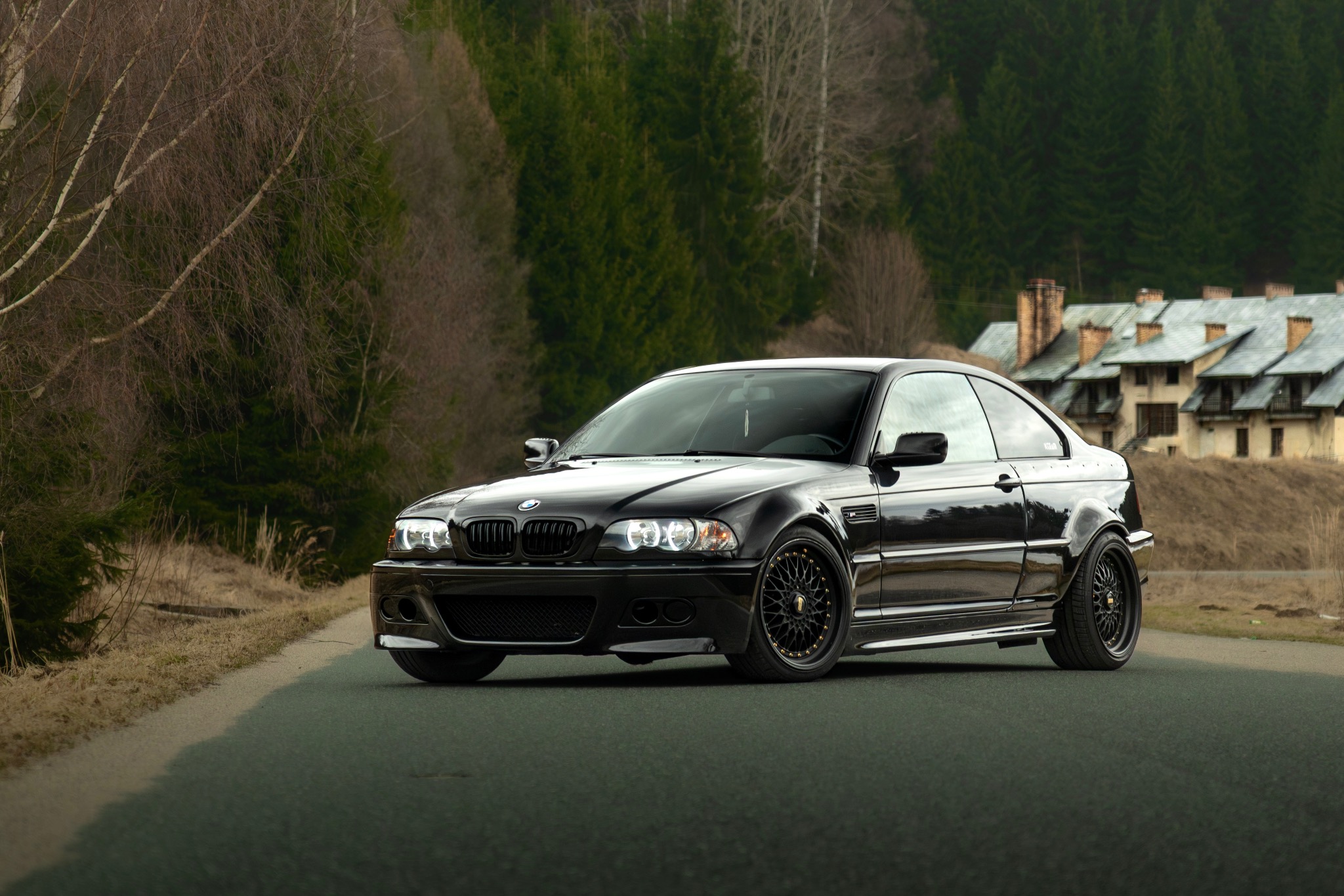 Czarna, zmodyfikowana sportowa limuzyna BMW E46 Coupe na tle leśnego krajobrazu i zabudowań. Auto z czarnymi felgami BBS i obniżonym zawieszeniem.