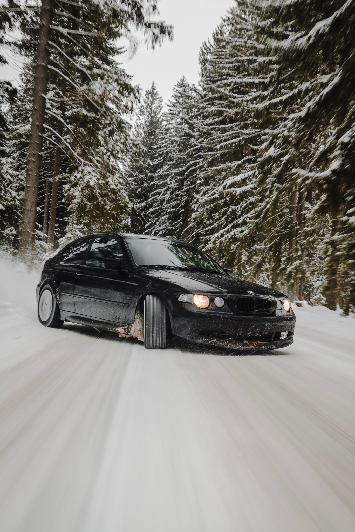 Dynamiczne ujęcie czarnego samochodu w zimowej scenerii Kłodzka. Rozbryzg śniegu spod kół, drzewa pokryte śniegiem w tle, ruch uchwycony w kadrze.