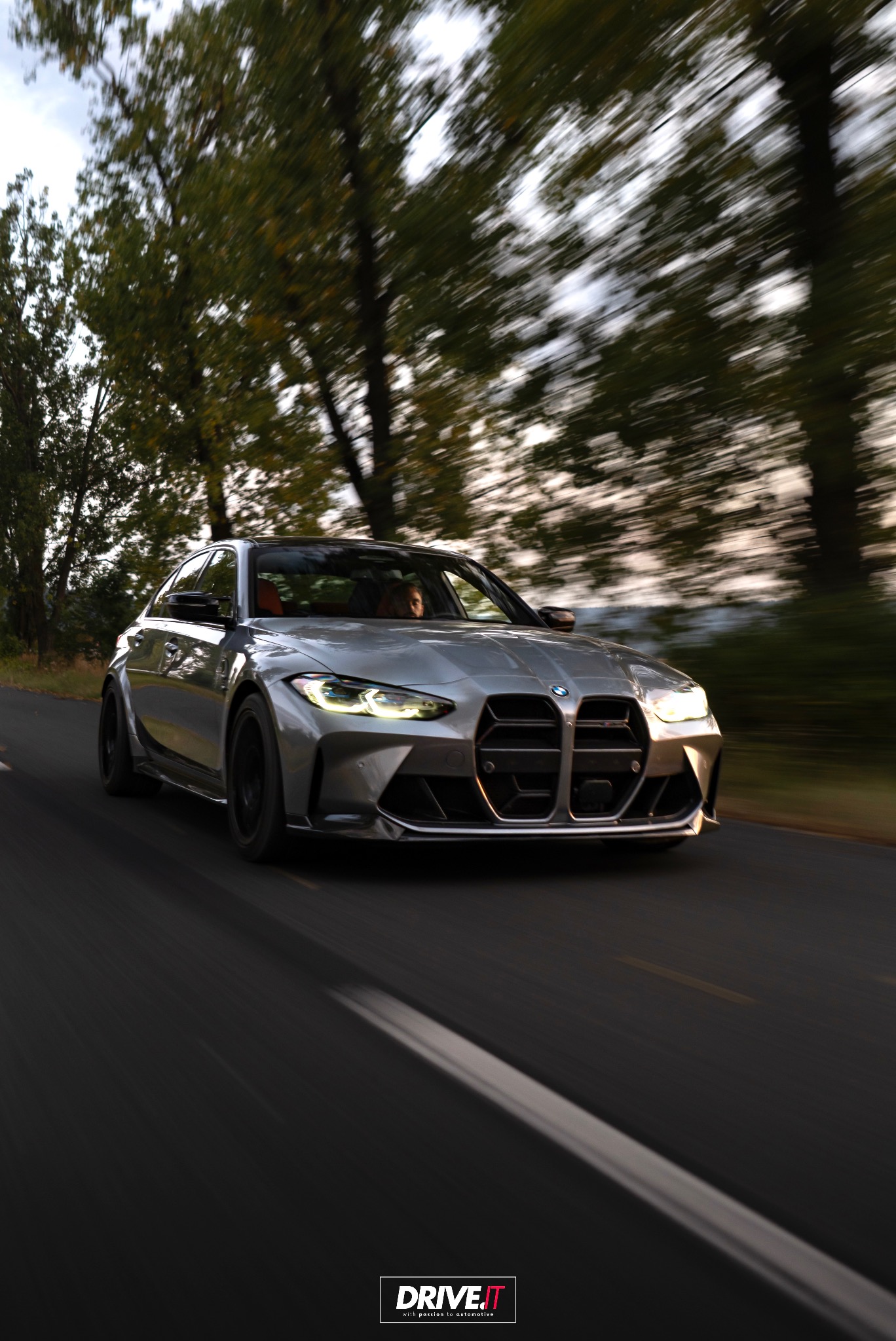 Dynamiczne ujęcie srebrnego BMW M4 w ruchu, z rozmytym tłem drzew i drogi, podkreślające prędkość i moc pojazdu. Widoczne detale karoserii i oświetlenia.