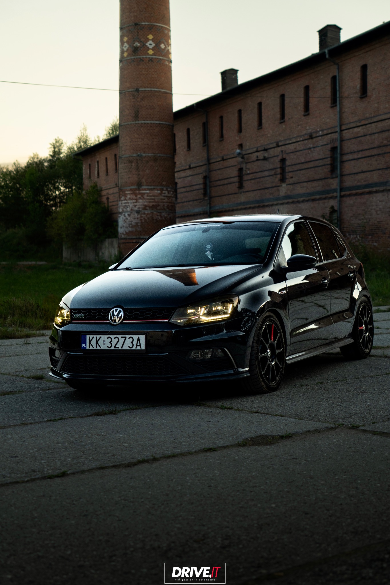Czarne auto VW Polo GTI na tle ceglanego budynku z kominem. Reflektory włączone, tablica rejestracyjna widoczna. Zdjęcie wykonane o zmierzchu.