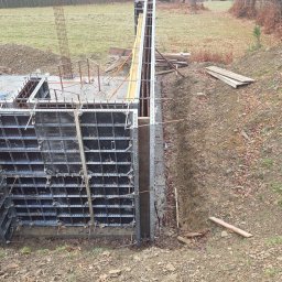 Eduard Benedzisiuk ŻELBET BUD - Szare szalunki fundamentowe z metalowymi elementami, widoczne zbrojenia i wykop, w tle łąka i las. Ujęcie z góry ukazuje etap budowy fundamentów.