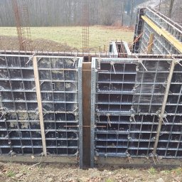 Eduard Benedzisiuk ŻELBET BUD - Szare szalunki fundamentowe zbrojone stalą, ustawione w ziemi na budowie. Widoczne drewniane podpory i elementy mocujące. W tle trawa, drzewa i pochmurne niebo.