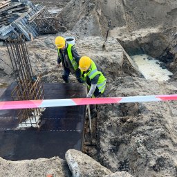 Eduard Benedzisiuk ŻELBET BUD - Dwóch robotników w kaskach i kamizelkach pracuje przy szalunku fundamentu zbrojonego, otoczonego wykopem i ziemią. Widoczne narzędzia i taśma ostrzegawcza.