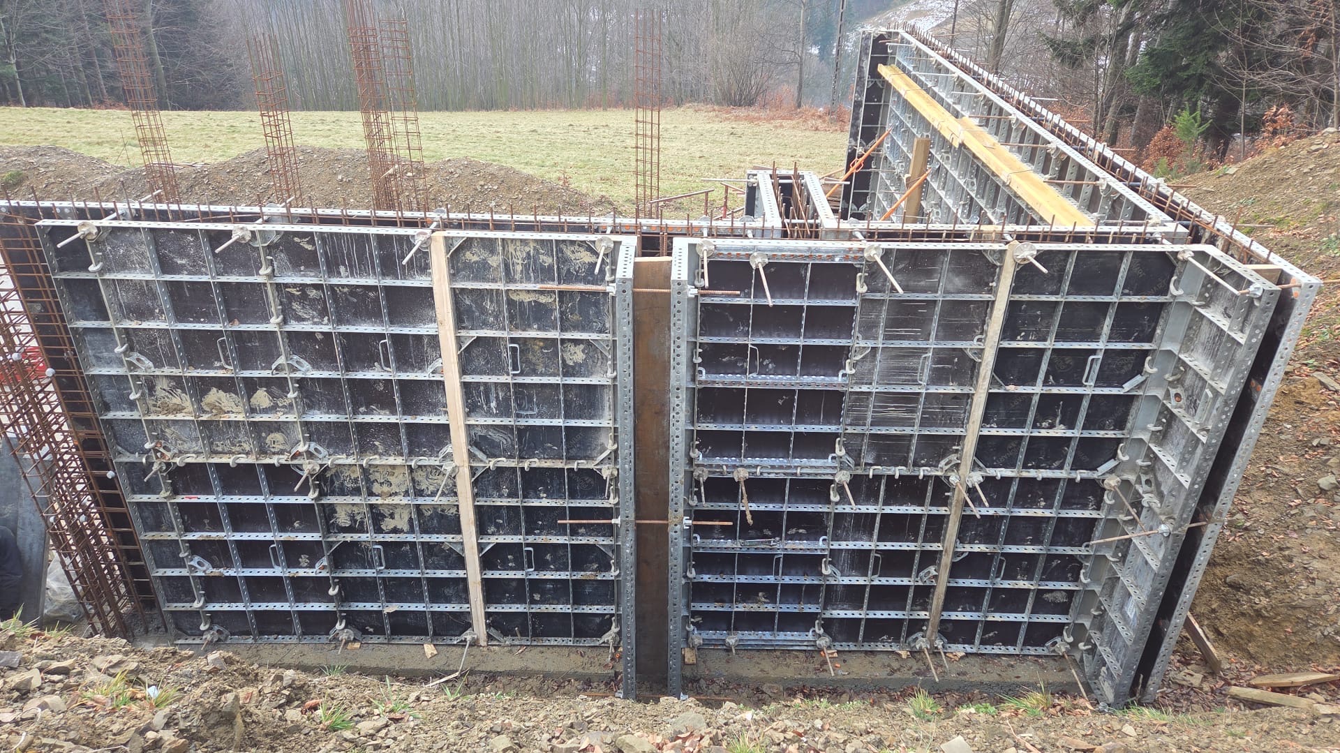 Szare szalunki fundamentowe zbrojone stalą, ustawione w ziemi na budowie. Widoczne drewniane podpory i elementy mocujące. W tle trawa, drzewa i pochmurne niebo.
