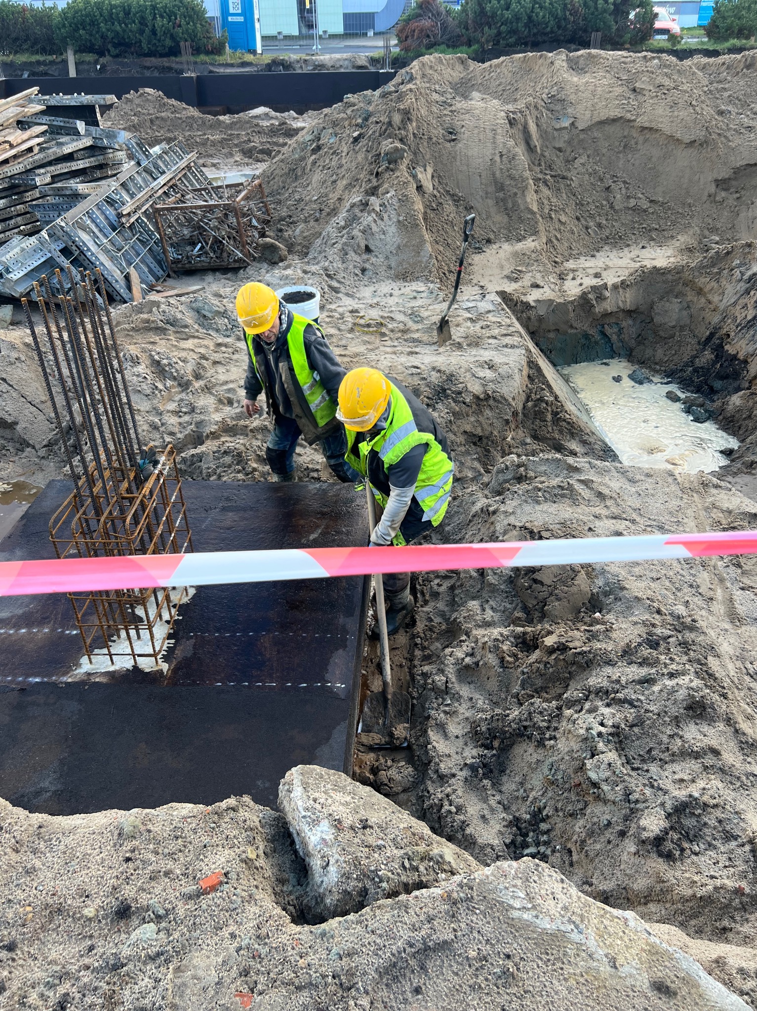 Dwóch robotników w kaskach i kamizelkach pracuje przy szalunku fundamentu zbrojonego, otoczonego wykopem i ziemią. Widoczne narzędzia i taśma ostrzegawcza.