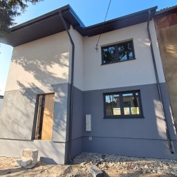 ŚWIST-BUD - Dwukondygnacyjny dom po ociepleniu elewacji, z szarą dolną i białą górną częścią. Widoczne czarne okna i drzwi wejściowe. Teren wokół domu w trakcie prac wykończeniowych.