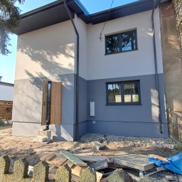 ŚWIST-BUD - Dwukondygnacyjny dom po remoncie elewacji, z otwartymi drzwiami wejściowymi i dwukolorową fasadą (szary dół, beżowa góra). Widoczne gruzy i ogrodzenie.