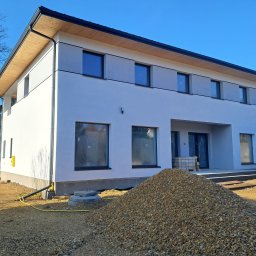 ŚWIST-BUD - Dwukondygnacyjny dom z jasną elewacją i drewnianym wykończeniem dachu, widoczny stos żwiru przed budynkiem i częściowo otwarty samochód dostawczy.