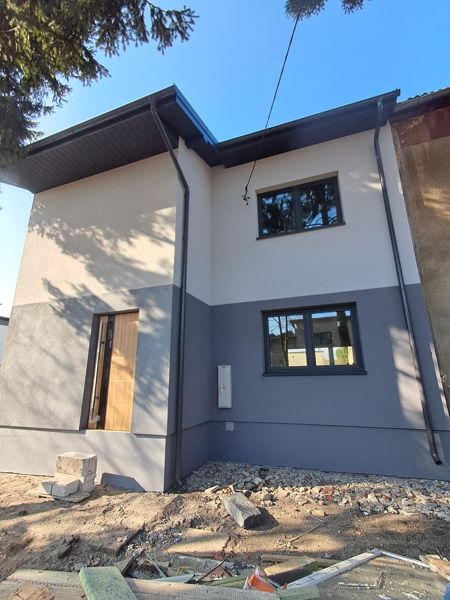 Dwukondygnacyjny dom po ociepleniu elewacji, z szarą dolną i białą górną częścią. Widoczne czarne okna i drzwi wejściowe. Teren wokół domu w trakcie prac wykończeniowych.