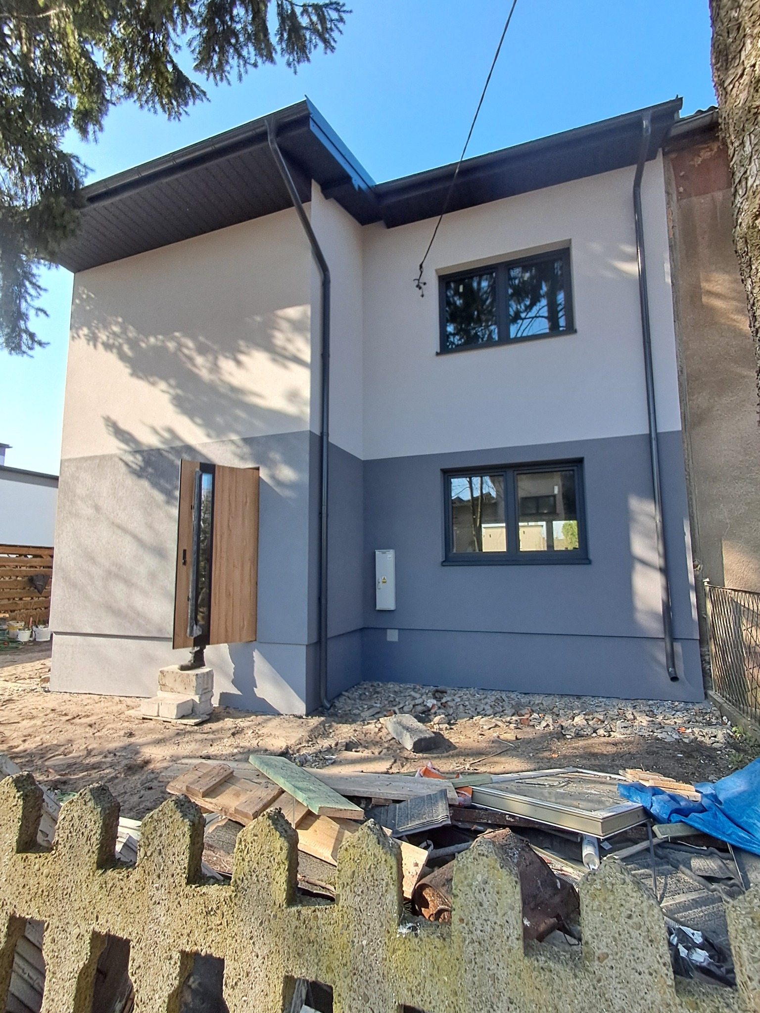 Dwukondygnacyjny dom po remoncie elewacji, z otwartymi drzwiami wejściowymi i dwukolorową fasadą (szary dół, beżowa góra). Widoczne gruzy i ogrodzenie.