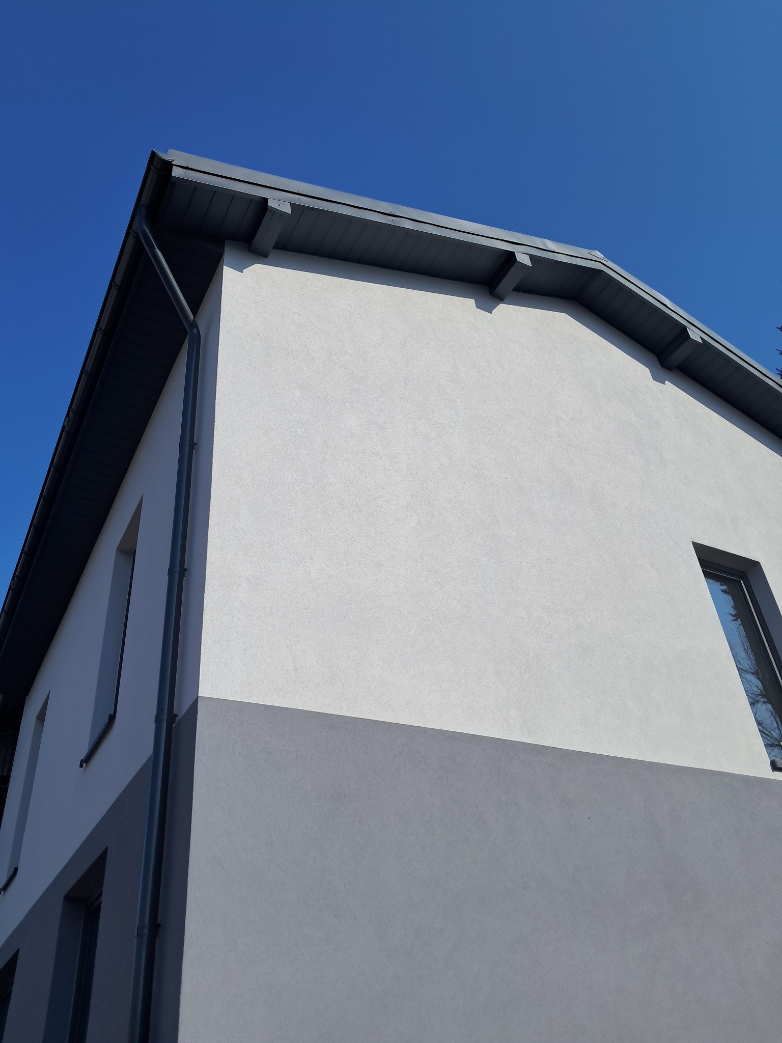 Dwukolorowa elewacja domu w odcieniach szarości i bieli, z widocznymi oknami i rynnami, sfotografowana pod kątem w słoneczny dzień.