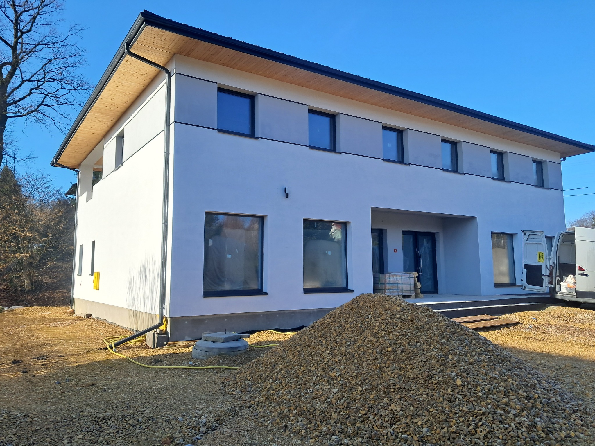 Dwukondygnacyjny dom z jasną elewacją i drewnianym wykończeniem dachu, widoczny stos żwiru przed budynkiem i częściowo otwarty samochód dostawczy.