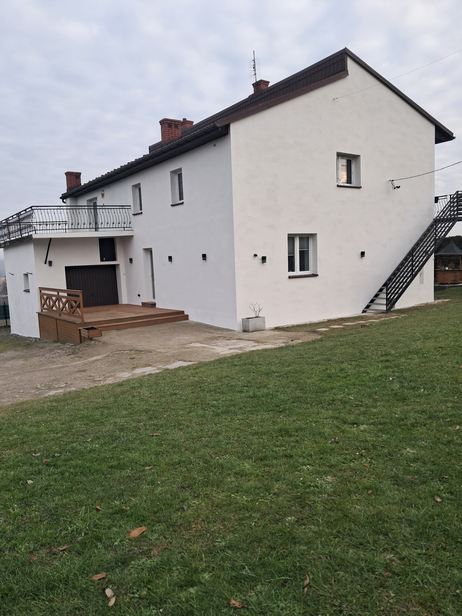 Biały dom z drewnianym tarasem i zewnętrznymi schodami, widok z trawnika. Nowoczesne oświetlenie elewacyjne. Ciemna dachówka i balustrada balkonu.