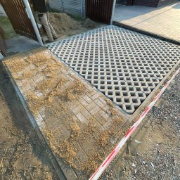 Mateusz Witek - Fragment podjazdu z ażurowych płyt betonowych i kostki, częściowo zasypanej piaskiem. W tle widoczne ogrodzenie posesji i osoba przy bramie.