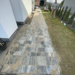 Mateusz Witek - Ścieżka z kostki brukowej w trakcie spoinowania piaskiem, widok z góry. W tle ogrodzenie panelowe i osoba. Nowoczesne budownictwo jednorodzinne.