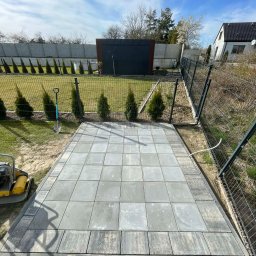 Mateusz Witek - Ułożona, szara kostka brukowa na przygotowanym podłożu, widoczna ubijarka, szpadel i fragment ogrodzenia panelowego. Tło: trawnik, tuje i budynek.