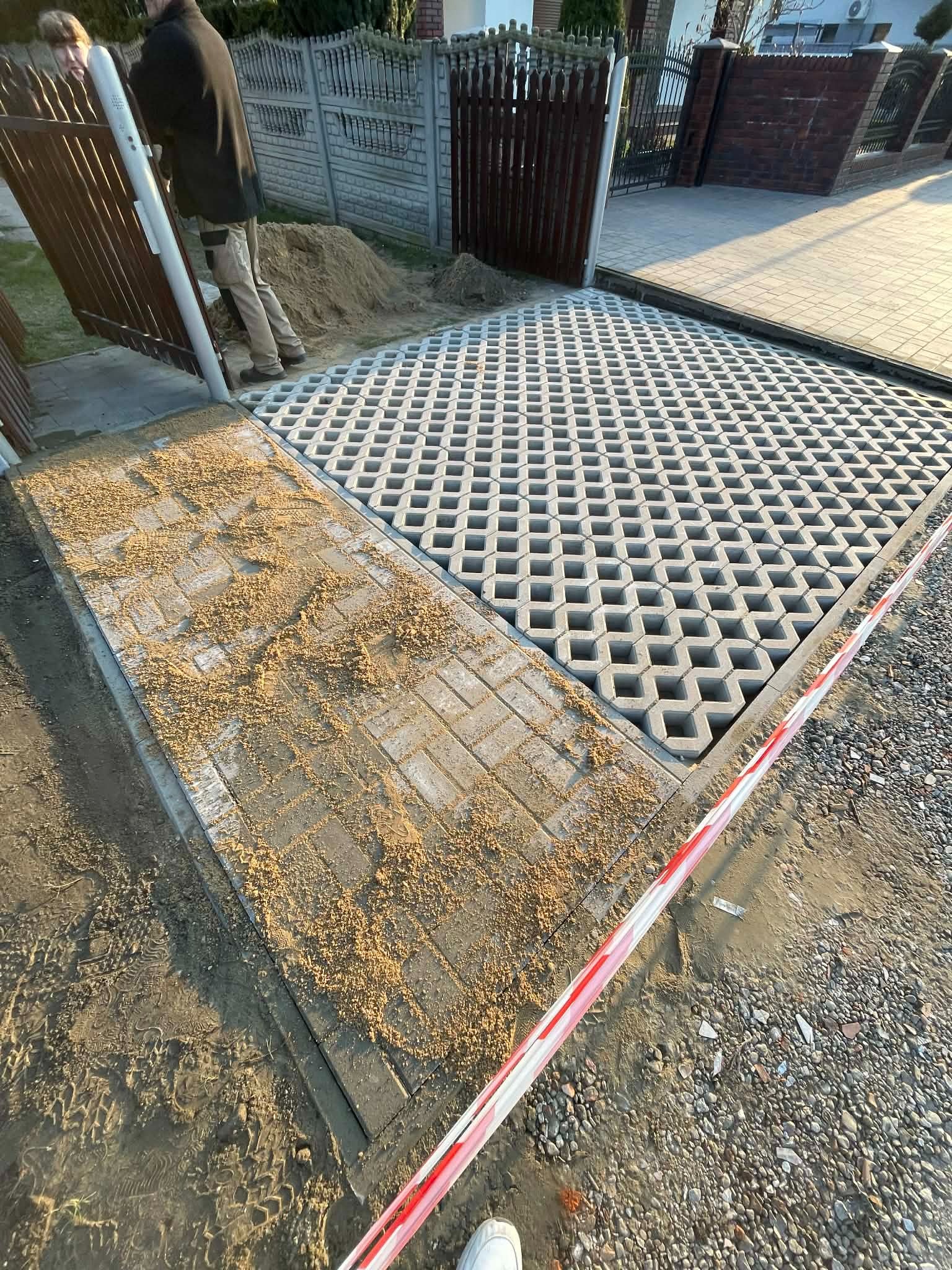 Fragment podjazdu z ażurowych płyt betonowych i kostki, częściowo zasypanej piaskiem. W tle widoczne ogrodzenie posesji i osoba przy bramie.
