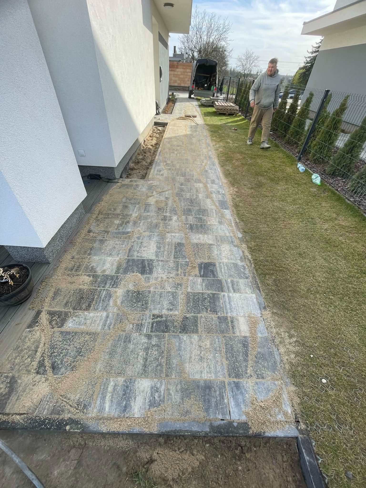 Ścieżka z kostki brukowej w trakcie spoinowania piaskiem, widok z góry. W tle ogrodzenie panelowe i osoba. Nowoczesne budownictwo jednorodzinne.