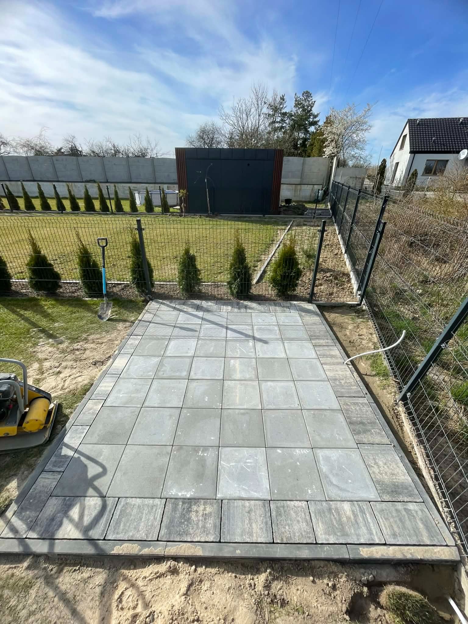Ułożona, szara kostka brukowa na przygotowanym podłożu, widoczna ubijarka, szpadel i fragment ogrodzenia panelowego. Tło: trawnik, tuje i budynek.