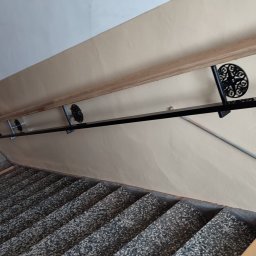 Blistal - Klatka schodowa z balustradą: drewniany poręcz i czarne, metalowe wsporniki z dekoracyjnymi rozetami. Widok z góry na schody pokryte lastryko.