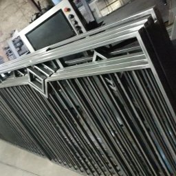Blistal - Stos srebrnych, metalowych elementów konstrukcyjnych z geometrycznym wzorem, ułożonych w warsztacie, z widocznym panelem sterowania maszyny w tle.