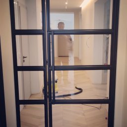Blistal - Nowoczesne, czarne drzwi wewnętrzne z metalową ramą i szklanymi panelami, widok na korytarz z parkietem w jodełkę i odkurzaczem. Minimalistyczny design.