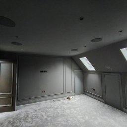 Prodecopainting - Pokój na poddaszu po remoncie: szare ściany z dekoracyjnymi listwami, okna dachowe, szara wykładzina, wbudowane głośniki sufitowe. Widoczne gniazdka elektryczne.