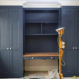 Prodecopainting - Granatowa zabudowa meblowa z biurkiem i półkami, oświetlona lampą budowlaną. Widoczne ślady prac wykończeniowych na podłodze.