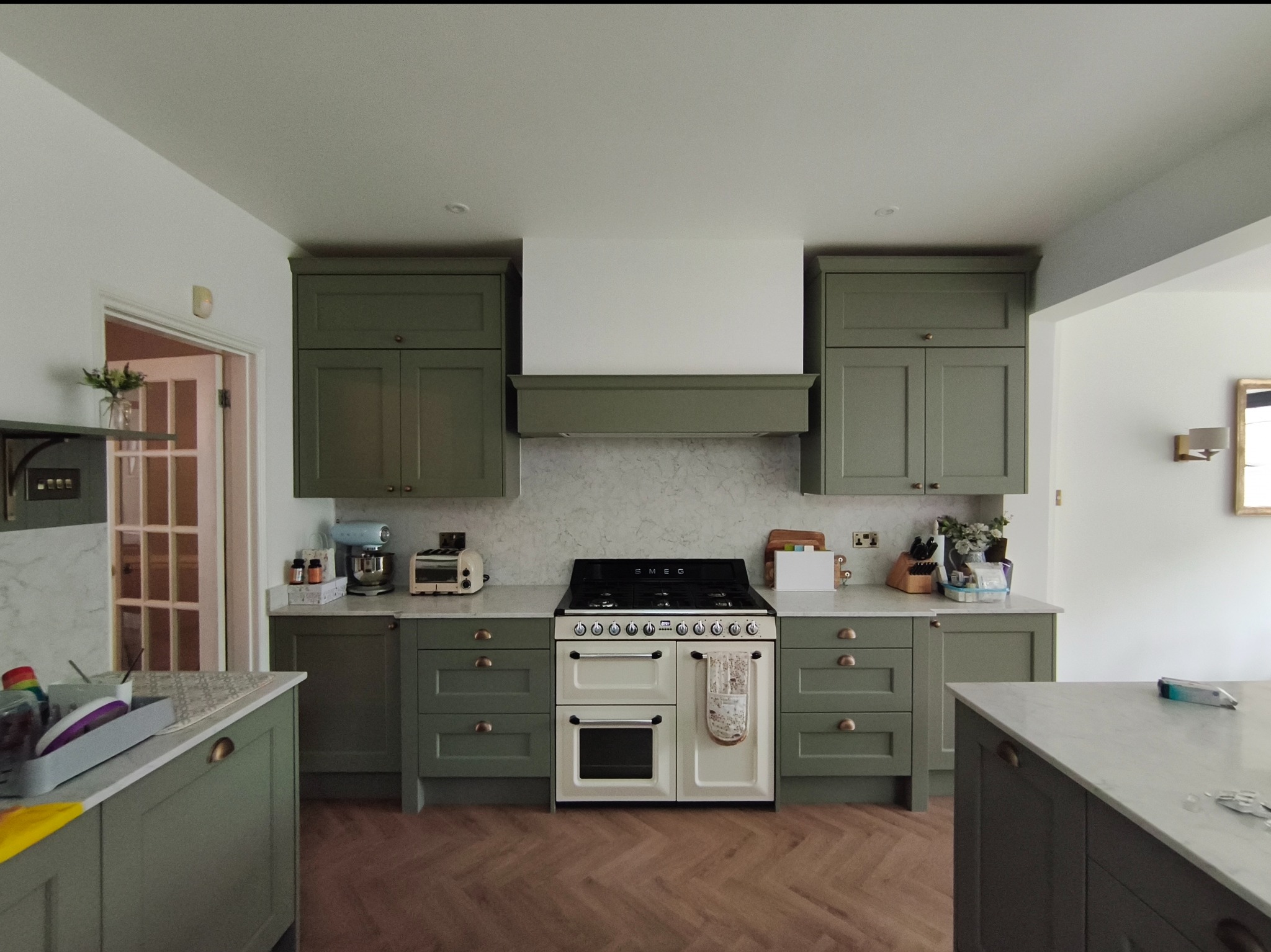 Kuchnia w odcieniach zieleni z drewnianą podłogą w jodełkę. Biały blat i sprzęt AGD. Widok na wyspę kuchenną i otwarte przejście do innego pomieszczenia.