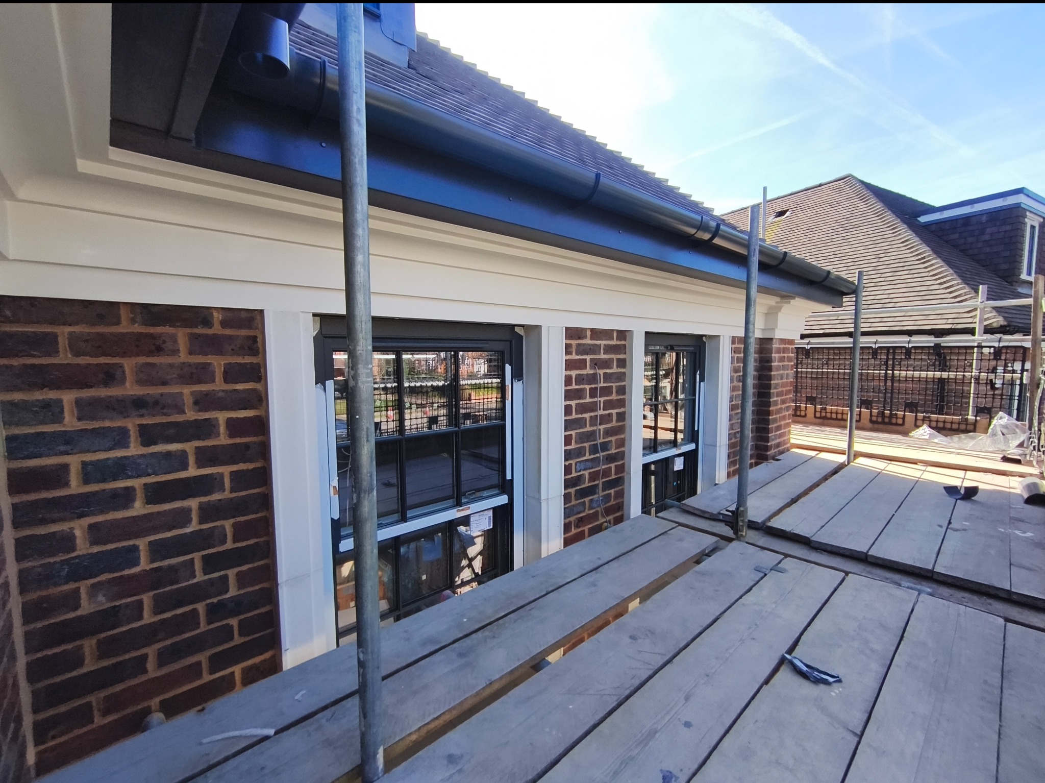 Elewacja budynku z cegły po renowacji, widok z rusztowania. Nowe okna z czarnymi ramami, białe obramowania i czarne rynny. Słoneczny dzień.