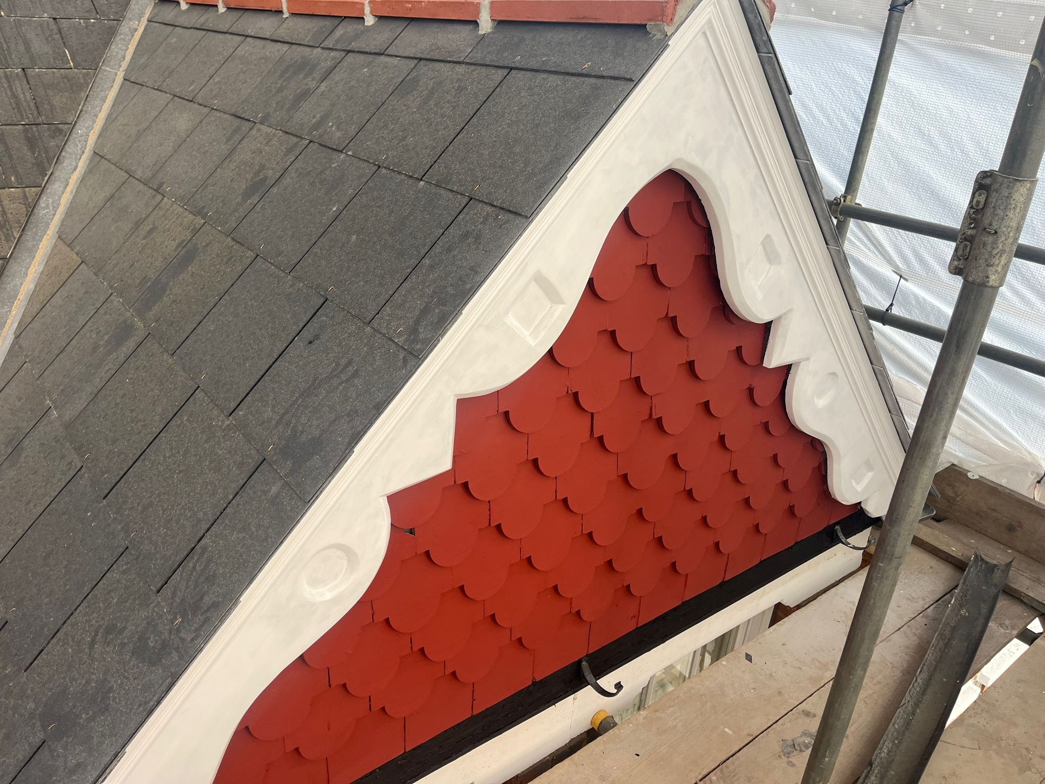 Fragment dachu z dekoracyjnym wykończeniem w kolorze czerwonym i białym, widoczne rusztowanie. Nietypowe ułożenie dachówki.