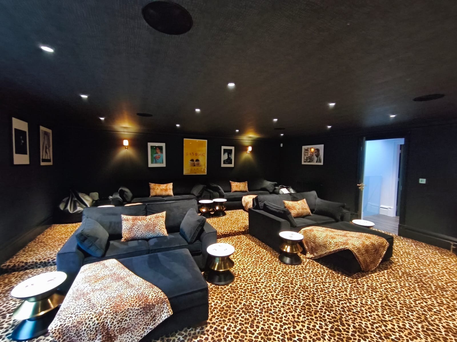 Czarne ściany i sufit w kinie domowym z sofami i dywanem w panterkę. Oświetlenie punktowe i kinkiety. Plakaty na ścianach. Widok na otwarte drzwi do innego pomieszczenia.
